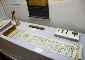 小尾権三郎の父から山田家に贈られた遺品。祈誓文が書かれた巻物（手前）や祈祷に用いる金銅製の法具（右奥の左側３点）などがある＝北杜市郷土資料館